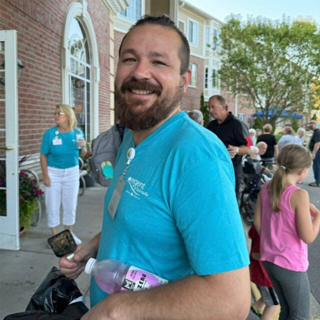 Chef Ricco, the director of culinary services at Regent at Burnsville, smiles while holding a drink and a grilling tool during The Regent's summer party. He is wearing a teal Regent-branded shirt, with a name badge visible. In the background, other attendees, including children and staff, are enjoying the event near the entrance of the building.