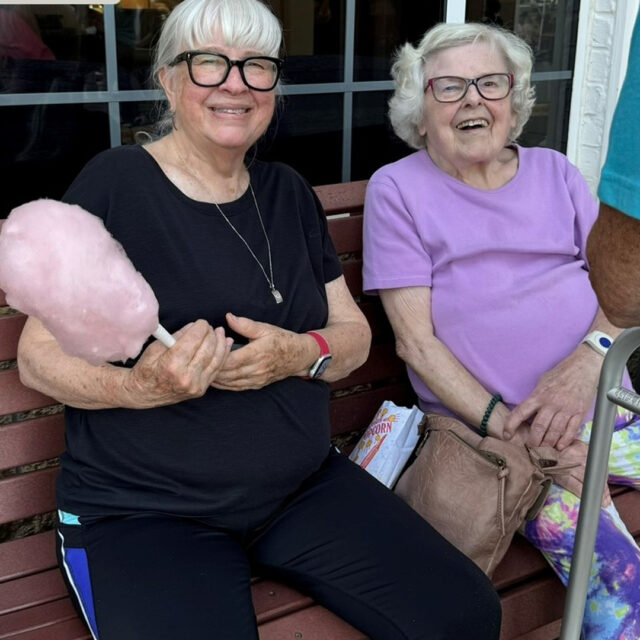 Two smiling women are sitting on a bench at The Regent's summer party. The woman on the left, wearing a black shirt and glasses, is holding pink cotton candy. The woman on the right, in a lavender shirt and glasses, is holding a purse with a popcorn bag beside her. Both are enjoying the festive atmosphere of the event.