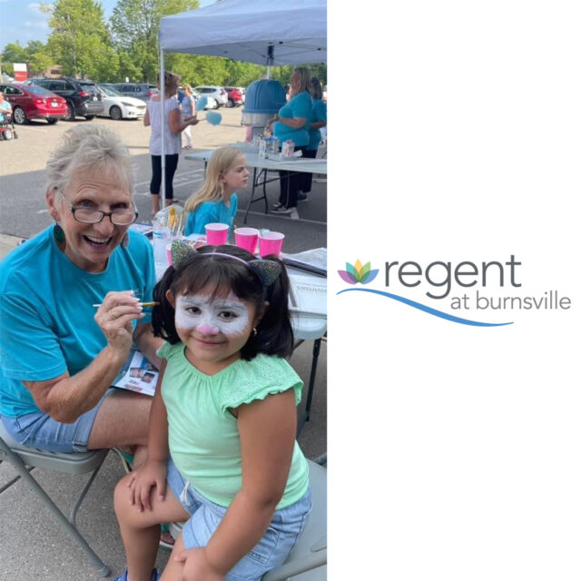 A cheerful woman in a teal t-shirt is painting the face of a young girl, who is smiling with her face painted.They are sitting outdoors under a tent at a community event. Other volunteers in teal shirts can be seen in the background, along with cotton candy supplies. The logo for Regent at Burnsville is displayed on the right side of the image.
