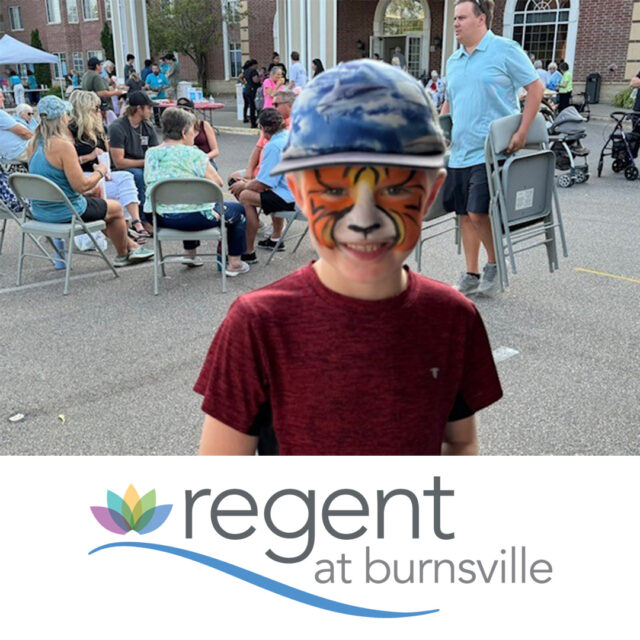 A smiling boy wearing a tiger face paint and a blue cap stands at The Regent's summer party. Behind him, attendees are seated and socializing outdoors near the entrance of the building. The festive atmosphere includes various activities and people enjoying the event. The Regent at Burnsville logo is displayed at the bottom of the image.