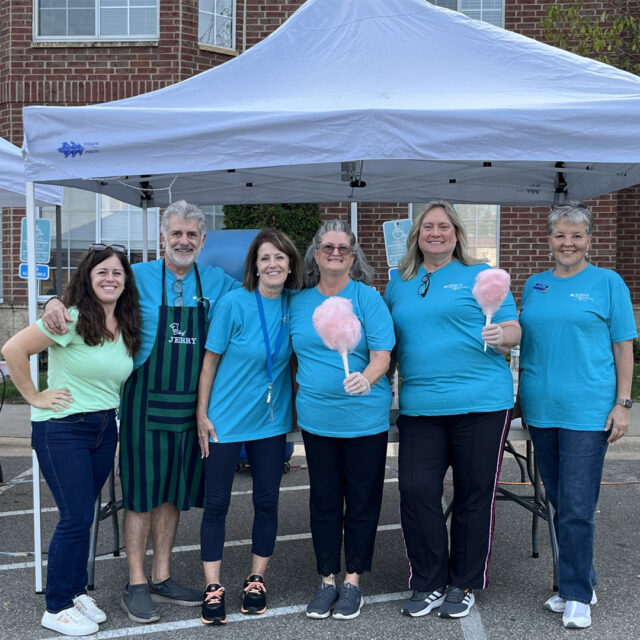 A group of six smiling volunteers is standing together under a tent at The Regent's summer party. Three of the women are holding pink cotton candy. They are all wearing teal shirts, except one woman on the far left in a light green top, and one man is wearing an apron. The brick building of The Regent is visible in the background, creating a warm and festive atmosphere at the event.