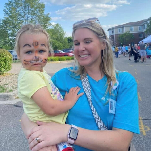 A smiling woman in a teal Regent shirt is holding a young child with face paint resembling a tiger at The Regent's summer party. The child, wearing a yellow shirt, looks curiously at the camera while the woman beams. The event is taking place outdoors with people, tents, and activities visible in the background, creating a lively and fun atmosphere.