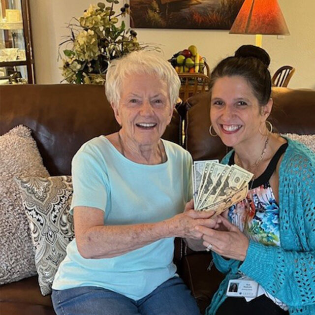 Director of sales and marketing, Sue Seykora, smiles as she presents $305 to lucky resident Nora, the winner of the split the pot raffle at The Regent's summer party. Nora, wearing a light blue shirt, holds the cash prize with a big smile. The other half of the raffle proceeds will go toward a special project at Regent at Burnsville. They are seated in a cozy living room with decorative pillows and plants in the background, creating a warm and joyful atmosphere.