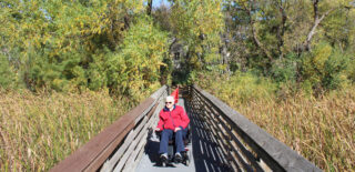 A resident of Regent at Burnsville enjoys a sunny day navigating the scenic Earley Lake Trail in a motorized chair, surrounded by vibrant greenery and peaceful nature.